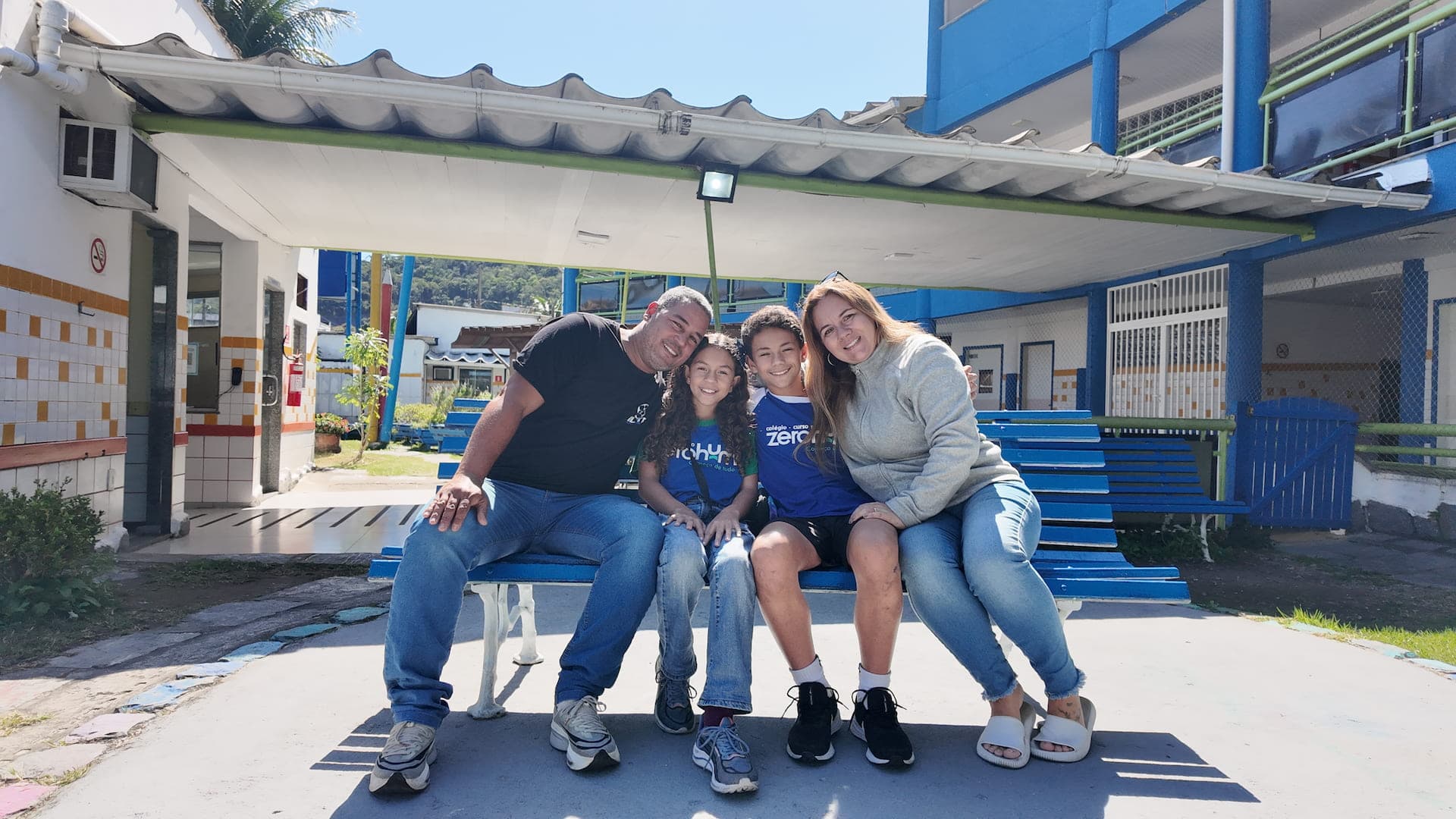 Pai, mãe e dois filhos pequenos de uniforme Zerohum, sentados num banco no pátio da escola
